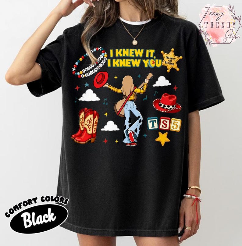 May include: A black t-shirt featuring a graphic print with the text I KNEW IT, I KNEW YOU, TS5, and INFINITY AND BEYOND. The design includes a central figure with a guitar, red cowboy boots, a red hat, a beaded bracelet, and colourful stars.