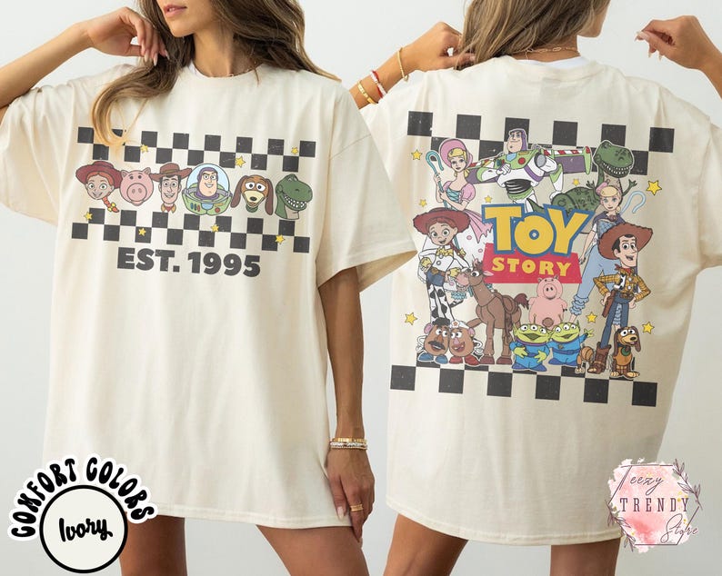 Op de afbeelding: Ivoor T-shirt met een dambordpatroon en Toy Story-stripfiguren. De voorkant heeft gezichten van personages en "EST. 1995". De achterkant toont meer personages en de woorden "Toy Story".