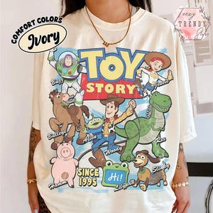May include: An ivory t-shirt featuring a graphic print of Toy Story characters including Buzz Lightyear, Jessie, Bullseye, Woody, Rex, Hamm, Slinky, and an Alien. The text reads Toy Story, Since 1995, and Hi, with character names labeled.