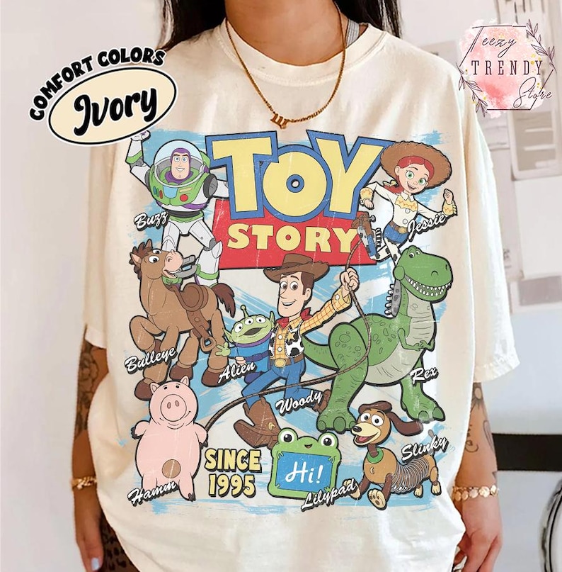May include: An ivory t-shirt featuring a graphic print of Toy Story characters including Buzz Lightyear, Jessie, Bullseye, Woody, Rex, Hamm, Slinky, and an Alien. The text reads Toy Story, Since 1995, and Hi, with character names labeled.