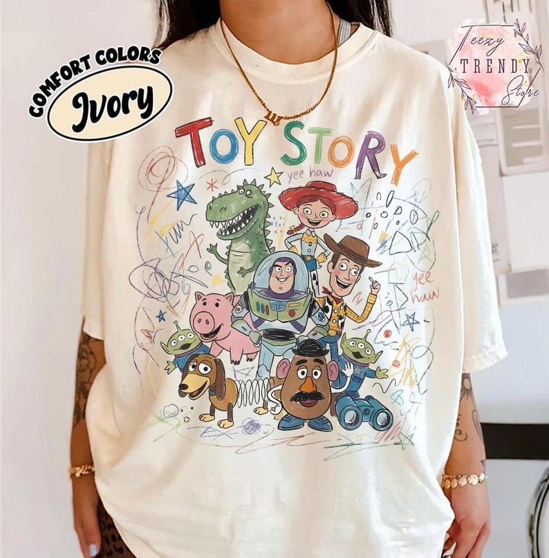 May include: Ivory-colored t-shirt featuring a colorful graphic of Toy Story characters. The design includes Woody, Buzz Lightyear, and other characters from the animated film. The words "Toy Story" are written in a playful font.