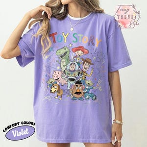 May include: Violet Comfort Colors t-shirt featuring a colorful graphic of Toy Story characters. The design includes Woody, Buzz Lightyear, and other beloved characters from the animated film. The shirt has short sleeves and a relaxed fit.