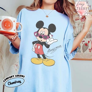 May include: Light blue t-shirt featuring a Mickey Mouse graphic wearing pink sunglasses. The cartoon character is in a confident pose with the words "Mickey Mouse" in a script font. The shirt has the words "Comfort Colors Chambray" on the bottom.
