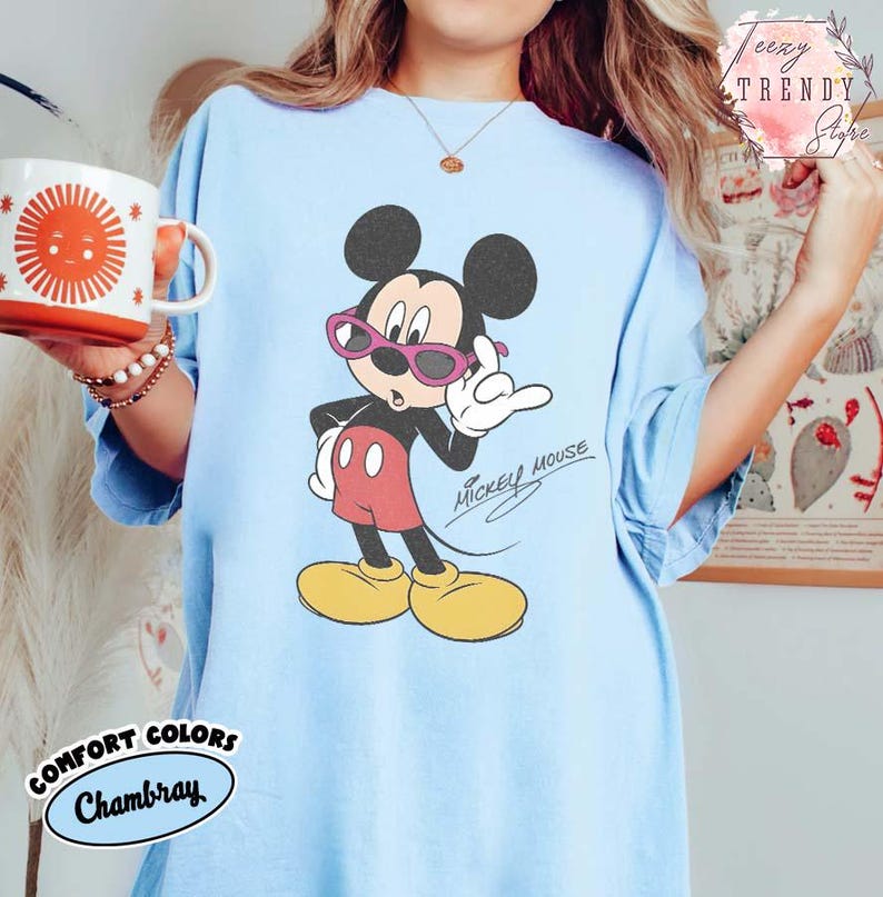 May include: Light blue t-shirt featuring a Mickey Mouse graphic wearing pink sunglasses. The cartoon character is in a confident pose with the words "Mickey Mouse" in a script font. The shirt has the words "Comfort Colors Chambray" on the bottom.