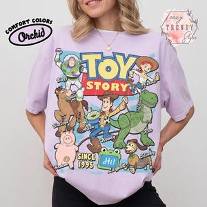 May include: A light purple t-shirt featuring a large graphic print of Toy Story characters including Buzz Lightyear, Jessie, Bullseye, Alien, Woody, Rex, Hamm, and Slinky Dog. The text reads Toy Story, Since 1995, and Hi on a green lilypad.