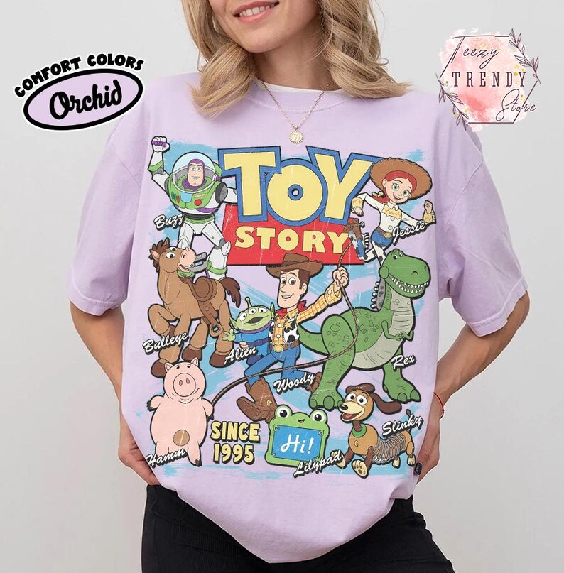 May include: A light purple t-shirt featuring a large graphic print of Toy Story characters including Buzz Lightyear, Jessie, Bullseye, Alien, Woody, Rex, Hamm, and Slinky Dog. The text reads Toy Story, Since 1995, and Hi on a green lilypad.