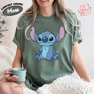 May include: Moss green t-shirt featuring a blue cartoon character with large ears and a friendly expression. The shirt has rolled-up sleeves and the words "Comfort Colors" and "Moss" are printed on the upper left. The brand name "Teezy Trendy Style" is printed on the upper right.