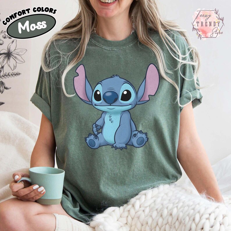 May include: Moss green t-shirt featuring a blue cartoon character with large ears and a friendly expression. The shirt has rolled-up sleeves and the words "Comfort Colors" and "Moss" are printed on the upper left. The brand name "Teezy Trendy Style" is printed on the upper right.
