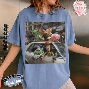 Puede incluir: Una camiseta azul de manga corta con un estampado gr&aacute;fico de los personajes de Toy Story, incluyendo a Woody, Buzz Lightyear, el Sr. Patata, Rex y Hamm. Los personajes aparecen en una escena cinematogr&aacute;fica desde un coche.