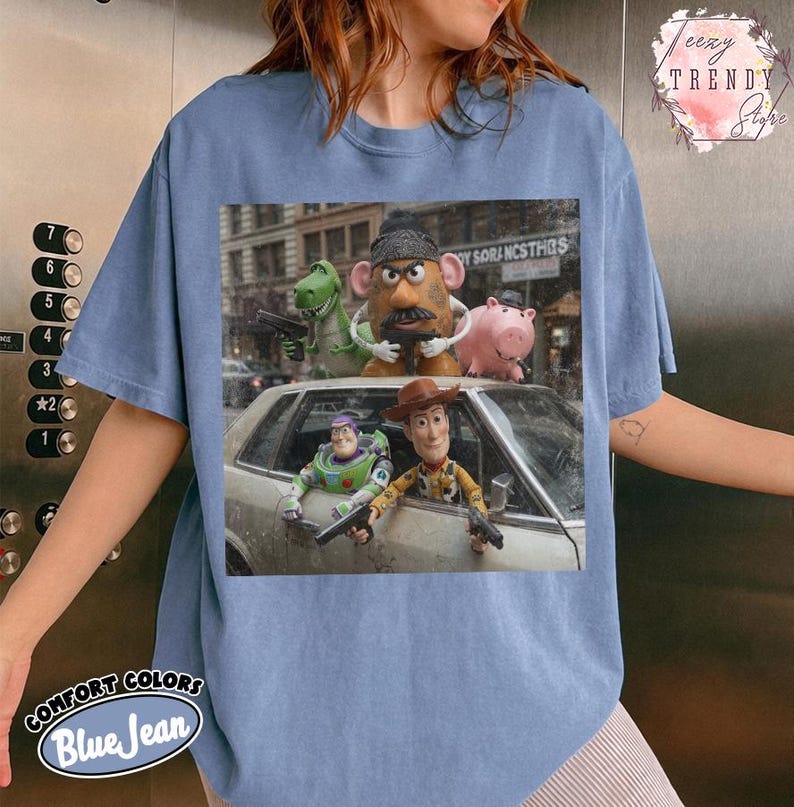 Puede incluir: Una camiseta azul de manga corta con un estampado gr&aacute;fico de los personajes de Toy Story, incluyendo a Woody, Buzz Lightyear, el Sr. Patata, Rex y Hamm. Los personajes aparecen en una escena cinematogr&aacute;fica desde un coche.