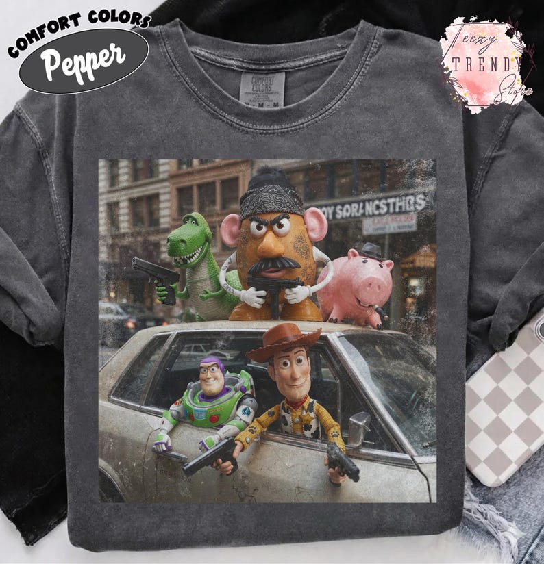 Puede incluir: Una camiseta de cuello redondo gris carb&oacute;n con un estampado gr&aacute;fico de cinco personajes animados de pel&iacute;cula, incluyendo un vaquero, un guardi&aacute;n espacial, una figura de patata, un dinosaurio verde y un cerdo rosa, sosteniendo pistolas de juguete negras.