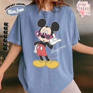 May include: Blue jean colored t-shirt featuring a graphic of Mickey Mouse wearing pink sunglasses. Mickey Mouse is in a pose with his hand up. The text "Mickey Mouse" is written in cursive. The shirt has the text "COMFORT COLORS" and "Blue Jean".
