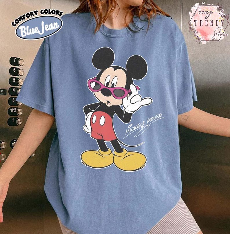 May include: Blue jean colored t-shirt featuring a graphic of Mickey Mouse wearing pink sunglasses. Mickey Mouse is in a pose with his hand up. The text "Mickey Mouse" is written in cursive. The shirt has the text "COMFORT COLORS" and "Blue Jean".