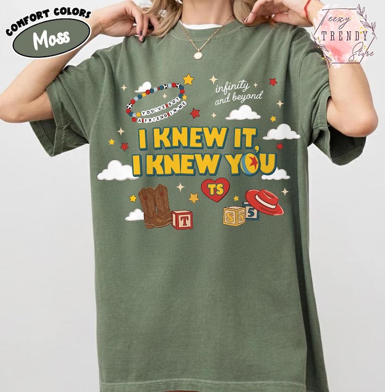 Puede incluir: Una camiseta de manga corta color verde musgo con un estampado gr&aacute;fico que incluye el texto "I KNEW IT, I KNEW YOU", "infinity and beyond" y "YOU'VE GOT A FRIEND IN ME". El dise&ntilde;o muestra botas de vaquero, un sombrero rojo y cubos.