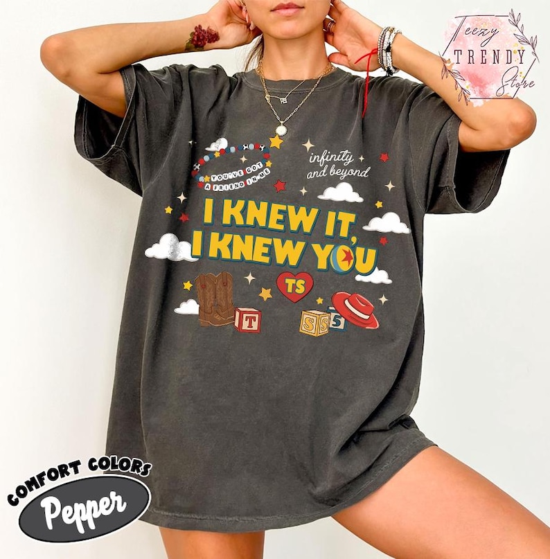 Puede incluir: Una camiseta de manga corta color gris carb&oacute;n con un estampado gr&aacute;fico que incluye el texto I KNEW IT, I KNEW YOU, infinity and beyond, y YOU'VE GOT A FRIEND IN ME. El dise&ntilde;o presenta ilustraciones de una bota, un sombrero y cubos.
