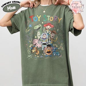 May include: Moss green t-shirt featuring the words "TOY STORY" in colorful letters with cartoon characters. The shirt has a crew neck and short sleeves. The words "Comfort Colors" are in a green oval on the upper left.