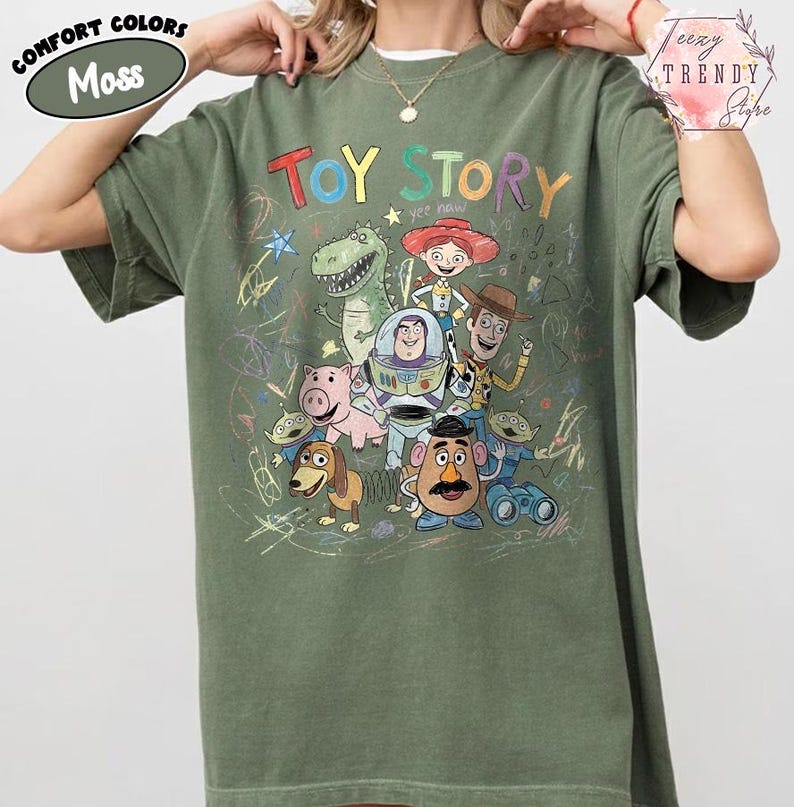May include: Moss green t-shirt featuring the words "TOY STORY" in colorful letters with cartoon characters. The shirt has a crew neck and short sleeves. The words "Comfort Colors" are in a green oval on the upper left.