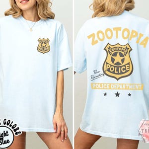 May include: Light blue t-shirt featuring a Zootopia Police badge on the front and back. The back of the shirt displays the words "ZOOTOPIA POLICE DEPARTMENT" in yellow, with "Now Recruiting" text. The shirt has a relaxed fit.
