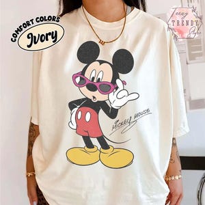 May include: Ivory-colored t-shirt featuring a graphic of Mickey Mouse wearing pink sunglasses. The cartoon character is in a pose with one hand raised. The words "Mickey Mouse" are written in cursive. The shirt also has the words "Comfort Colors" and "Ivory" in a circular design.