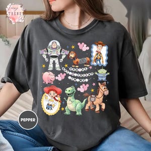 May include: Dark gray t-shirt featuring colorful cartoon characters from Toy Story. The design includes Buzz Lightyear, Woody, Jessie, and other characters, with the text "You've Got a Friend in Me."