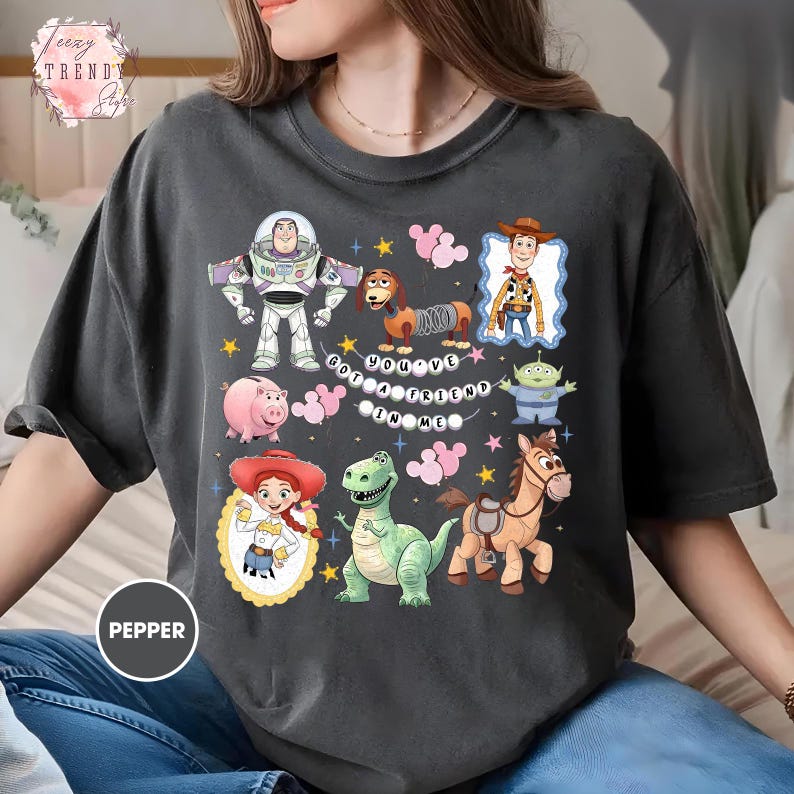 May include: Dark gray t-shirt featuring colorful cartoon characters from Toy Story. The design includes Buzz Lightyear, Woody, Jessie, and other characters, with the text "You've Got a Friend in Me."