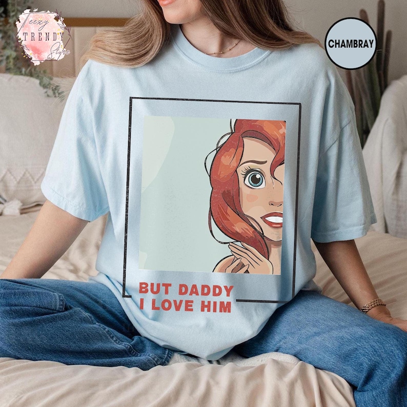 Puede incluir: Camiseta azul claro con una ilustraci&oacute;n enmarcada de un personaje pelirrojo y el texto "BUT DADDY I LOVE HIM". La camiseta tiene mangas cortas y cuello redondo. La camiseta est&aacute; hecha de un material suave.