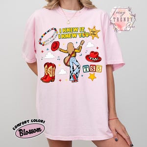 May include: A light pink t-shirt featuring a graphic print with the text I KNEW IT, I KNEW YOU, TS5, and INFINITY AND BEYOND. The design includes a person playing a guitar, a red cowboy hat, red cowboy boots, a beaded bracelet, and stars.