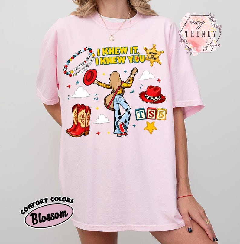 May include: A light pink t-shirt featuring a graphic print with the text I KNEW IT, I KNEW YOU, TS5, and INFINITY AND BEYOND. The design includes a person playing a guitar, a red cowboy hat, red cowboy boots, a beaded bracelet, and stars.