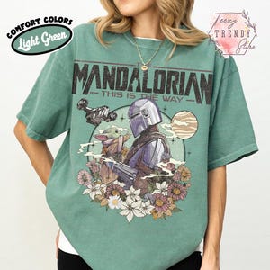 May include: Light green t-shirt with a graphic of The Mandalorian and Grogu, a spaceship, and the text "The Mandalorian - This is the Way." The design is surrounded by flowers. The shirt has the words "Comfort Colors" and "Light Green" on the upper left.