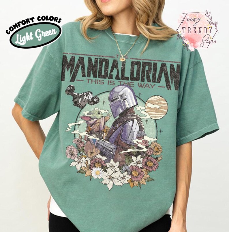 May include: Light green t-shirt with a graphic of The Mandalorian and Grogu, a spaceship, and the text "The Mandalorian - This is the Way." The design is surrounded by flowers. The shirt has the words "Comfort Colors" and "Light Green" on the upper left.