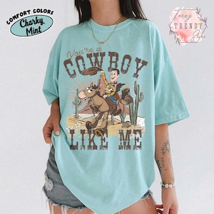 May include: A light mint green t-shirt featuring a graphic of a cowboy character riding a horse in a desert landscape with cacti. The text reads You're a COWBOY LIKE ME. The shirt includes branding text for Comfort Colors and Teezy Trendy Store.