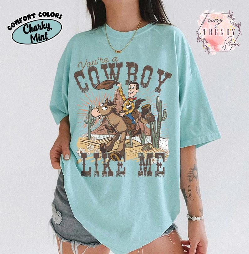 May include: A light mint green t-shirt featuring a graphic of a cowboy character riding a horse in a desert landscape with cacti. The text reads You're a COWBOY LIKE ME. The shirt includes branding text for Comfort Colors and Teezy Trendy Store.