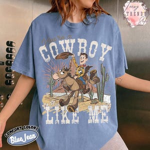 May include: A blue jean colored t-shirt featuring a graphic print of a cartoon cowboy riding a horse in a desert landscape with cacti. The text reads "You're a COWBOY LIKE ME" in a distressed white font. The design includes a brown hat and sunburst.