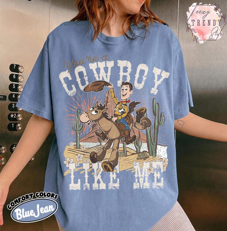 May include: A blue jean colored t-shirt featuring a graphic print of a cartoon cowboy riding a horse in a desert landscape with cacti. The text reads "You're a COWBOY LIKE ME" in a distressed white font. The design includes a brown hat and sunburst.