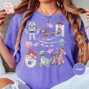 May include: A violet t-shirt featuring colorful illustrations of characters from the animated film "Toy Story." The design includes Buzz Lightyear, Woody, Jessie, Rex, Hamm, Slinky Dog, and other characters, with the phrase "You've Got a Friend in Me."