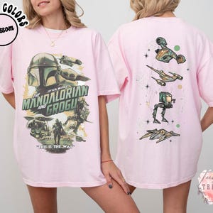 K&ouml;nnte beinhalten: Ein hellrosa T-Shirt mit einer Grafik von The Mandalorian und Grogu auf der Vorderseite, inklusive des Textes The Mandalorian and Grogu und This is the Way. Die R&uuml;ckseite zeigt eine vertikale Anordnung von vier Star Wars Fahrzeugen und Schiffen.
