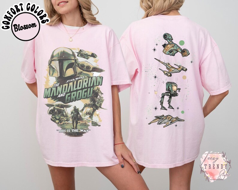 K&ouml;nnte beinhalten: Ein hellrosa T-Shirt mit einer Grafik von The Mandalorian und Grogu auf der Vorderseite, inklusive des Textes The Mandalorian and Grogu und This is the Way. Die R&uuml;ckseite zeigt eine vertikale Anordnung von vier Star Wars Fahrzeugen und Schiffen.