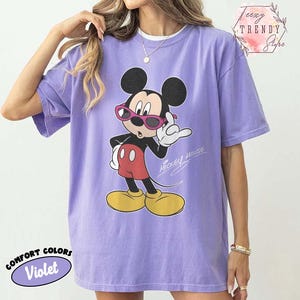 May include: Violet Comfort Colors t-shirt featuring a graphic of Mickey Mouse wearing pink sunglasses. Mickey Mouse is in a pose with one hand up. The words "Mickey Mouse" are written in script. The shirt also has the words "Comfort Colors" and "Violet" on it.