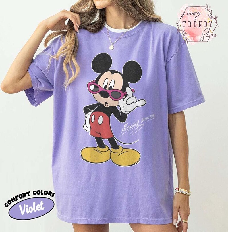 May include: Violet Comfort Colors t-shirt featuring a graphic of Mickey Mouse wearing pink sunglasses. Mickey Mouse is in a pose with one hand up. The words "Mickey Mouse" are written in script. The shirt also has the words "Comfort Colors" and "Violet" on it.