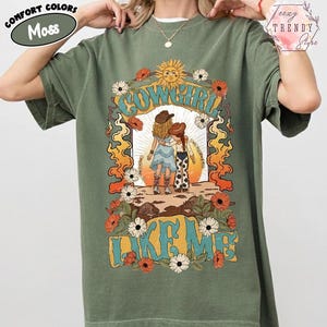 May include: A moss green short-sleeve t-shirt featuring a graphic print of two figures in cowboy hats and boots. The design includes the text "COWGIRL LIKE ME" surrounded by orange flames, white and red flowers, and a yellow sun in the background.