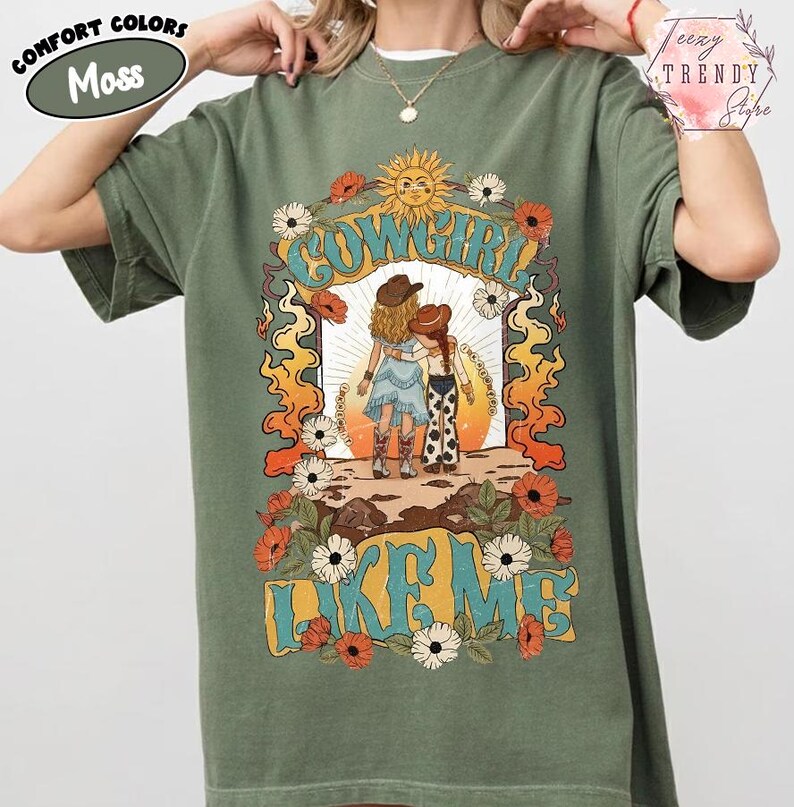 May include: A moss green short-sleeve t-shirt featuring a graphic print of two figures in cowboy hats and boots. The design includes the text "COWGIRL LIKE ME" surrounded by orange flames, white and red flowers, and a yellow sun in the background.