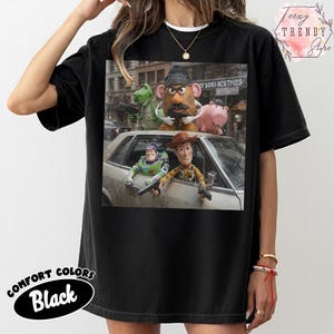 Puede incluir: Una camiseta negra con un estampado gr&aacute;fico de personajes de Toy Story, incluidos Woody, Buzz Lightyear, Mr. Potato Head, Rex y Hamm, posando con pistolas de juguete frente a un coche. El dise&ntilde;o muestra una escena cinematogr&aacute;fica con los personajes.