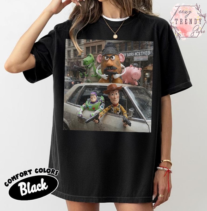Puede incluir: Una camiseta negra con un estampado gr&aacute;fico de personajes de Toy Story, incluidos Woody, Buzz Lightyear, Mr. Potato Head, Rex y Hamm, posando con pistolas de juguete frente a un coche. El dise&ntilde;o muestra una escena cinematogr&aacute;fica con los personajes.