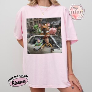 Puede incluir: Una camiseta rosa claro con un estampado gr&aacute;fico de personajes de Toy Story, incluidos Woody, Buzz Lightyear, Rex, Mr. Potato Head y Hamm, colocados dentro y sobre un coche. La camiseta incluye el texto Teezy Trendy Store.