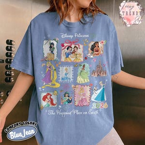 May include: Blue jean-colored t-shirt featuring a Disney Princess design. The design includes various princess illustrations, the text "2026", and the phrase "The Happiest Place on Earth". The shirt is from Comfort Colors.