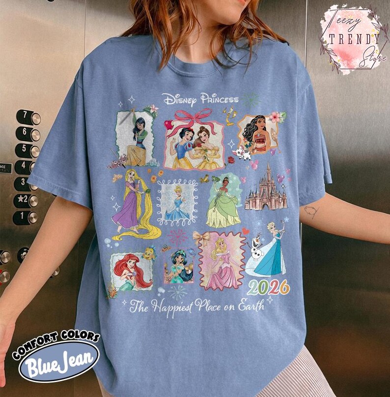 May include: Blue jean-colored t-shirt featuring a Disney Princess design. The design includes various princess illustrations, the text "2026", and the phrase "The Happiest Place on Earth". The shirt is from Comfort Colors.