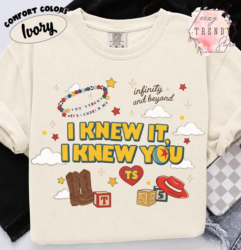 Puede incluir: Una camiseta de manga corta color marfil con un estampado gr&aacute;fico que incluye el texto "I KNEW IT, I KNEW YOU", "infinity and beyond" y "YOU'VE GOT A FRIEND IN ME". El dise&ntilde;o presenta ilustraciones de botas vaqueras, un sombrero rojo, bloques y estrellas.