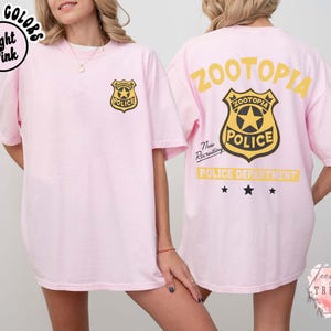 May include: Light pink t-shirt with a Zootopia Police badge on the front and back. The back of the shirt features the words "Zootopia Police Department" in yellow. The shirt is a comfort colors style.