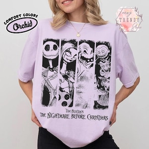 May include: A light purple t-shirt featuring a black graphic print of characters from Tim Burton's The Nightmare Before Christmas. The design includes Jack Skellington, Sally, Oogie Boogie, and other figures arranged in vertical panels with text below.
