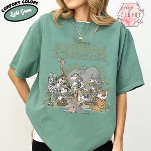 May include: Light green t-shirt with the text "Disney's Animal Kingdom" and a graphic of Disney characters and animals. The shirt has short sleeves and a crew neck. The words "Comfort Colors" and "Light Green" are in a green oval on the top left.